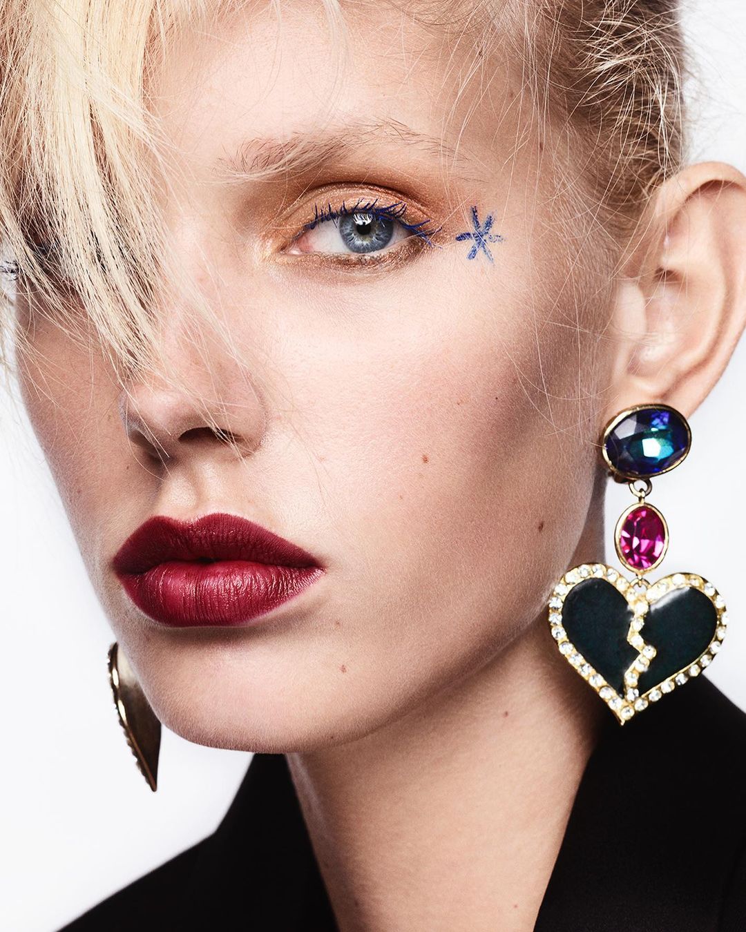 Close-up of a woman with blond hair, dark red lipstick, and a large, dangling heart-shaped earring. The shot is against a white background.