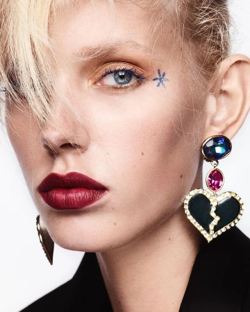 Close-up of a woman with blond hair, dark red lipstick, and a large, dangling heart-shaped earring. The shot is against a white background.