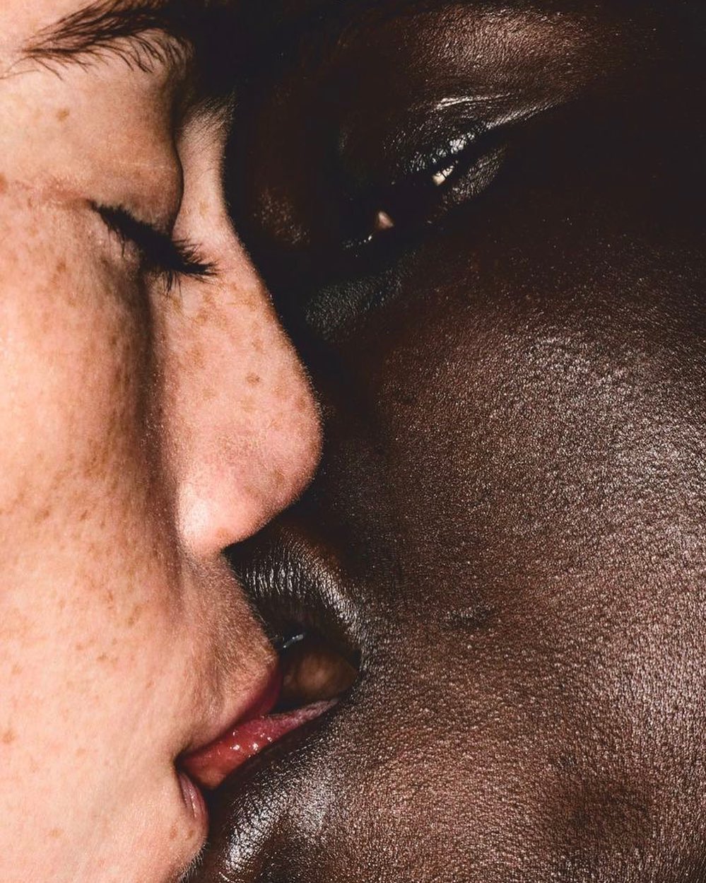 Close-up portrait of two faces of different skin tones, one light with freckles and one dark, very close together, almost kissing.
