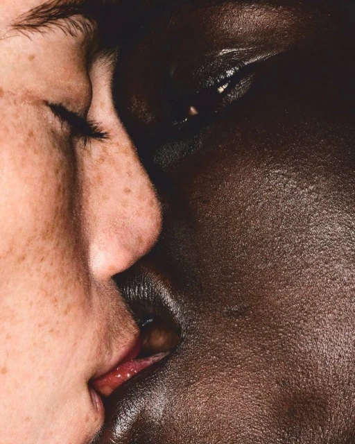 Close-up portrait of two faces of different skin tones, one light with freckles and one dark, very close together, almost kissing.