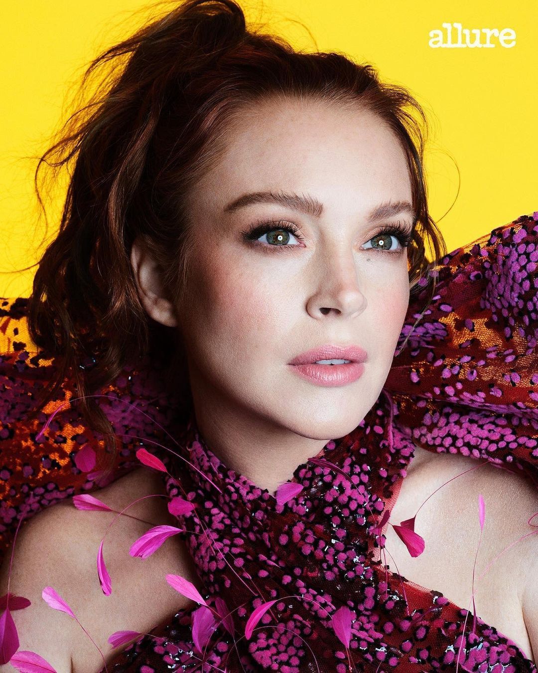 Close-up portrait of a beautiful adult woman with auburn hair, wearing a sequined, patterned dress and pink feathers against yellow backdrop.