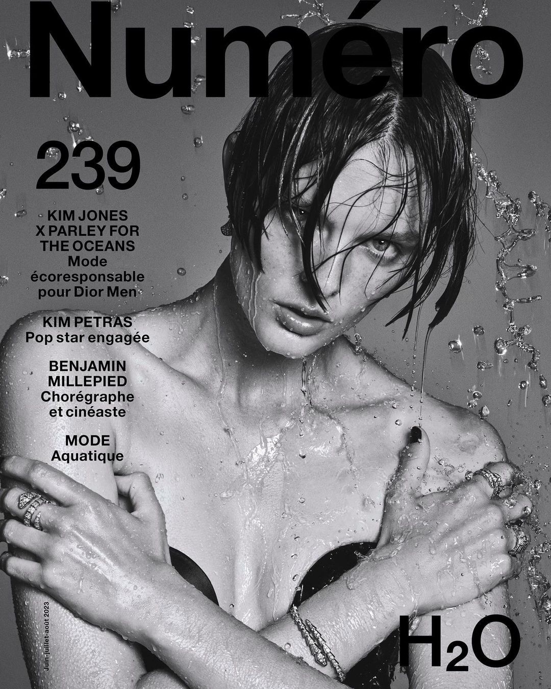 Black and white magazine cover featuring a model with wet hair and water droplets, adorned with jewelry and minimal clothing, H2O text.