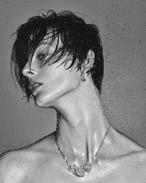 Monochromatic portrait of a shirtless person with wet dark hair and a jeweled necklace, facing left against a backdrop of water droplets.