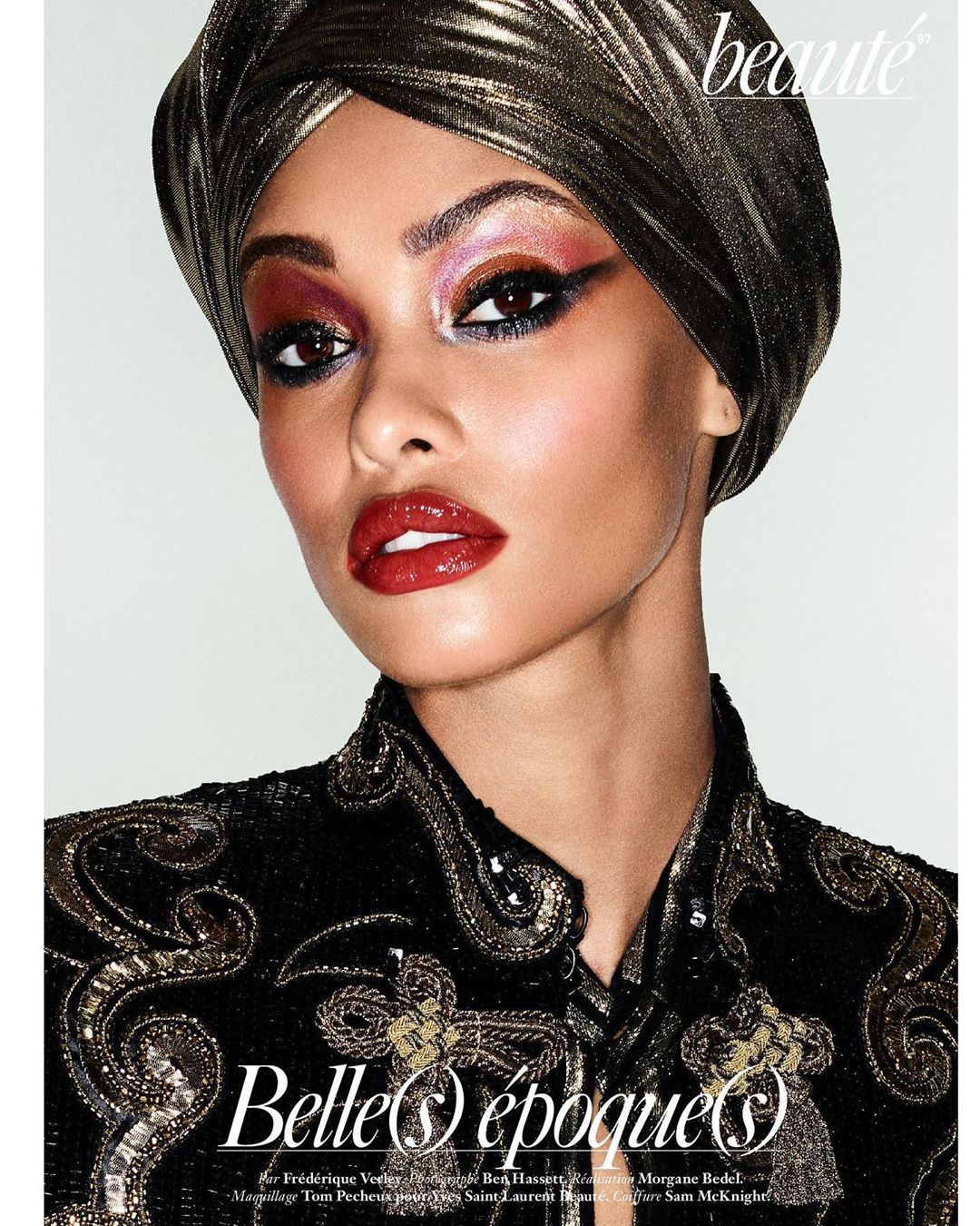 Glamorous model with dramatic eye makeup and glossy lips wears a gilded turban and beaded top against a clean backdrop.