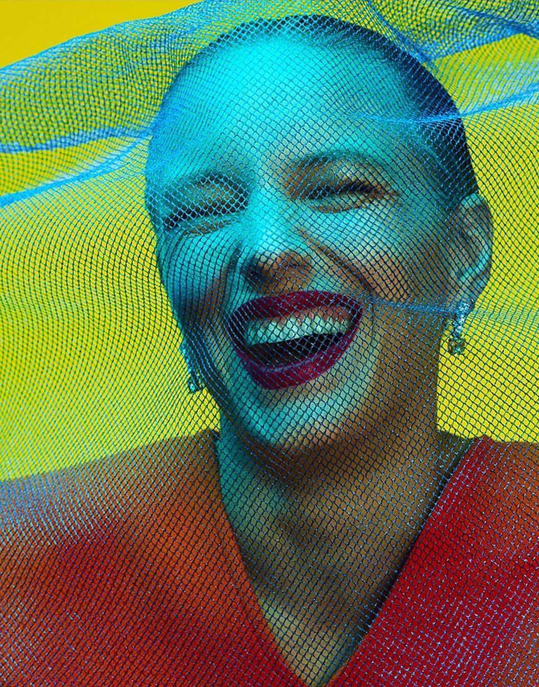 Smiling woman with eyes closed in orange top and blue mesh veil against bright yellow background in a portrait composition.