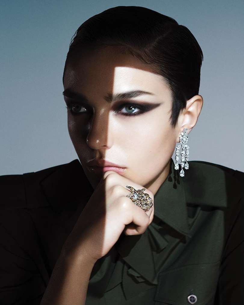 Close-up portrait of a woman with dramatic dark eye makeup, statement earrings, and a dark green shirt by Ben Hassett.
