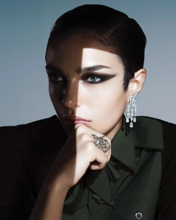 Close-up portrait of a woman with dramatic dark eye makeup, statement earrings, and a dark green shirt by Ben Hassett.