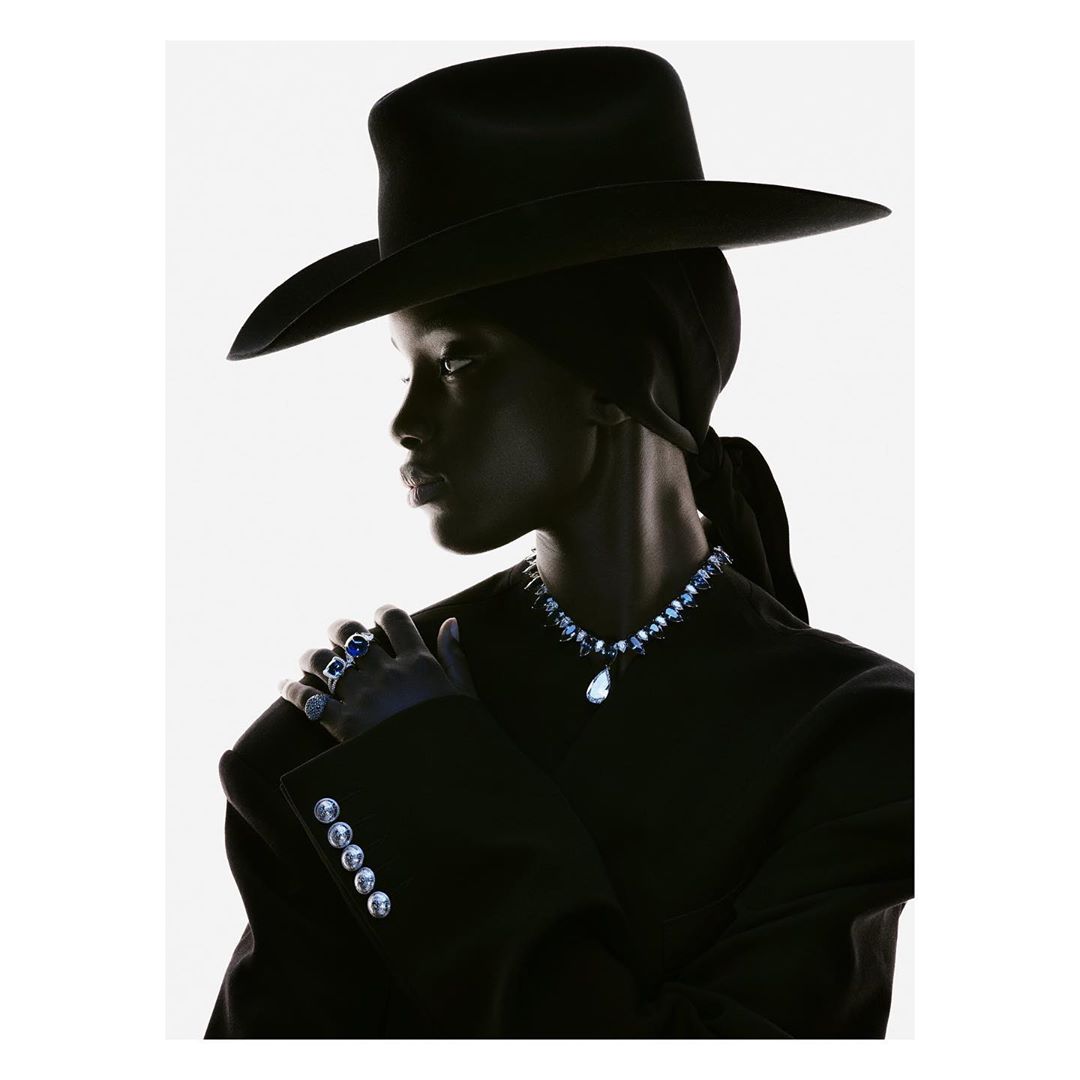 Dark-skinned model in black hat and jacket poses in high fashion studio portrait. Jewelry is visible. White background, high contrast.