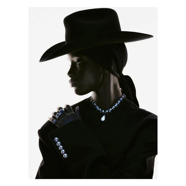 Dark-skinned model in black hat and jacket poses in high fashion studio portrait. Jewelry is visible. White background, high contrast.