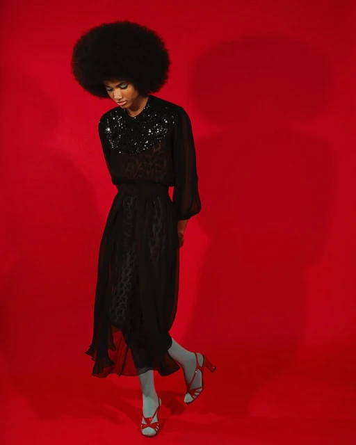 Elegant woman with afro in black dress and red heels poses against vibrant red background in a fashion forward studio portrait.