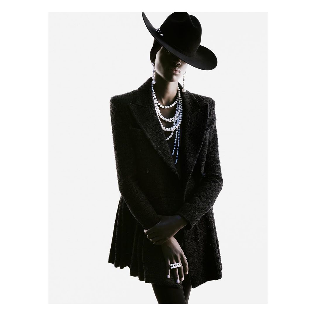 Fashion portrait of woman with pearl jewelry wearing black hat and coat in studio setting with bright, white background, looking at camera.