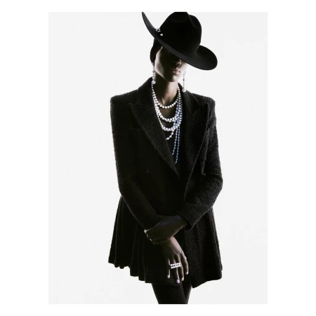 Fashion portrait of woman with pearl jewelry wearing black hat and coat in studio setting with bright, white background, looking at camera.