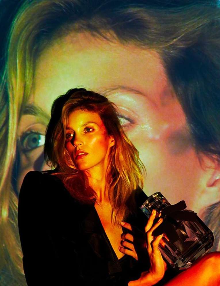 Adult female holding perfume bottle in a photo studio with filtered lighting and a facial portrait as a backdrop for a beauty advertisement.