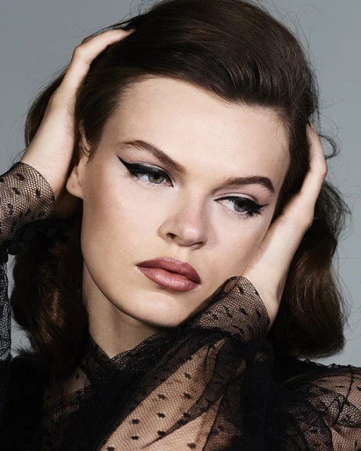 Close-up portrait of a young adult woman with dark hair and winged eyeliner, hands near her face, wearing a black dotted mesh top, against grey.