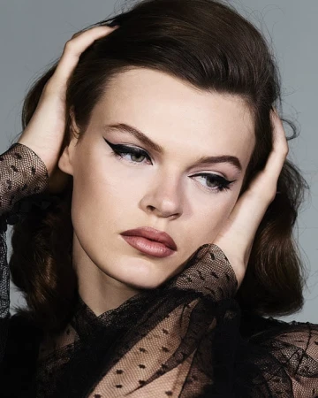 Close-up portrait of a young adult woman with dark hair and winged eyeliner, hands near her face, wearing a black dotted mesh top, against grey.