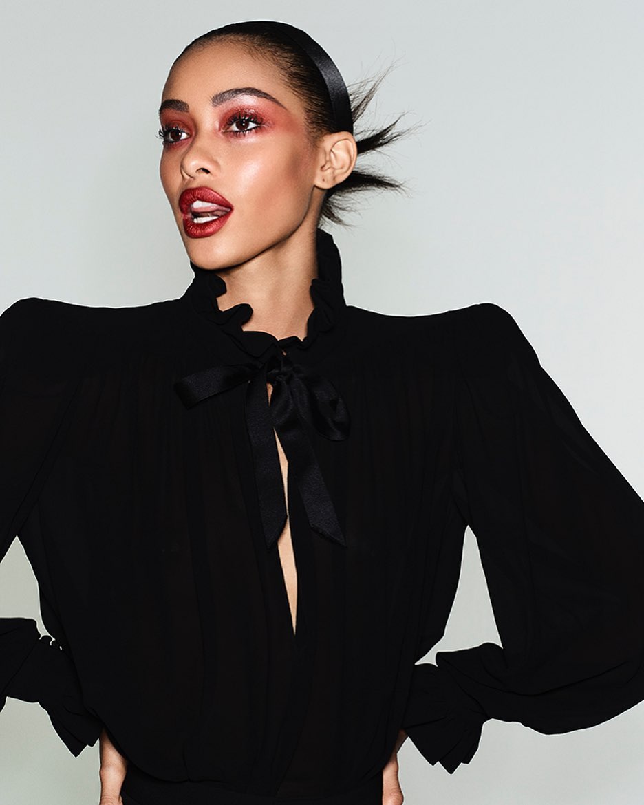 Close-up of a fashionable adult female with bold eye makeup and dark lipstick, posing in a black ruffled top against a plain background.