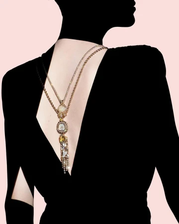 Back view of a person wearing a black dress and an ornate gold necklace with crystals on a light pink background, fashion photography.