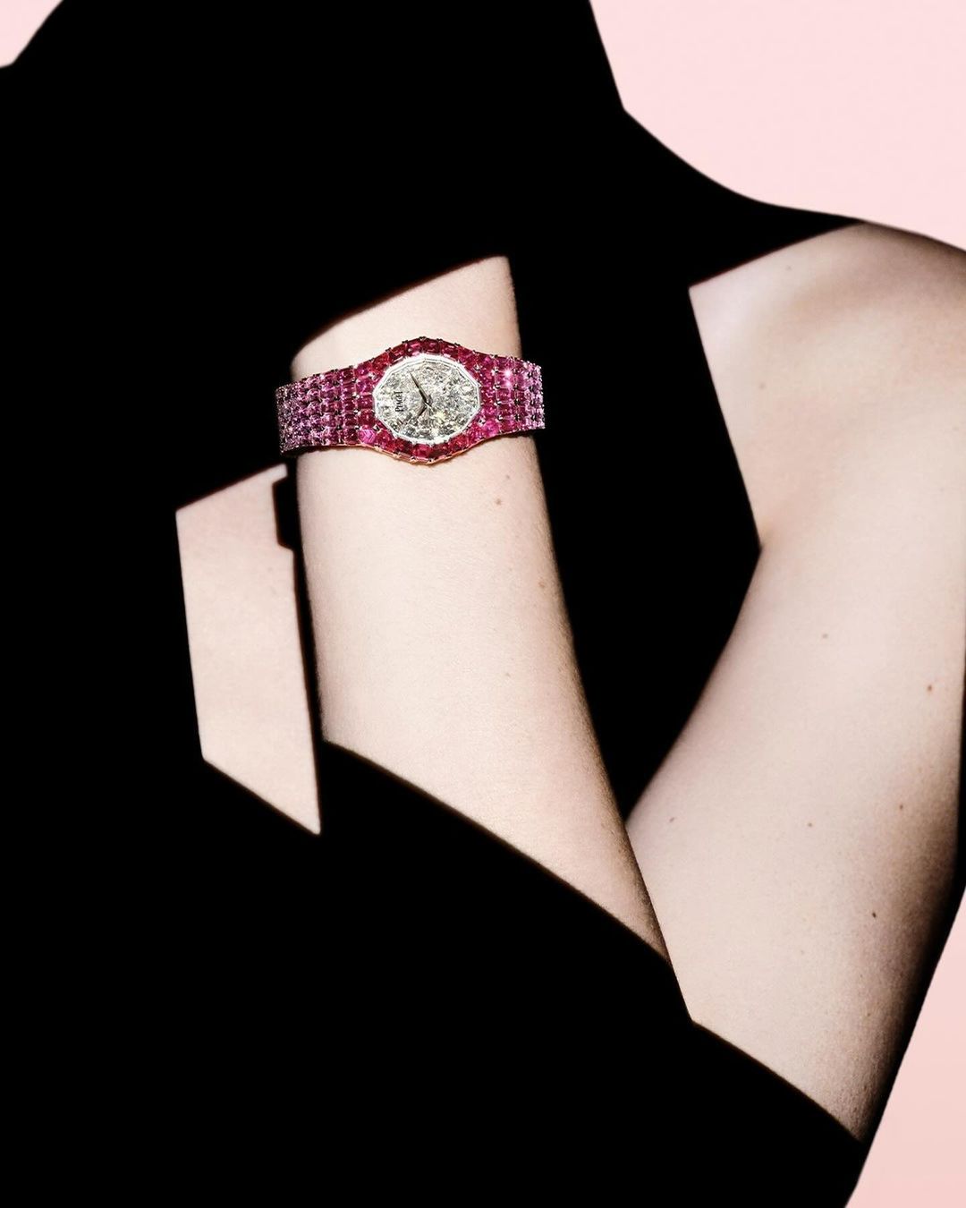 Close-up of a wrist wearing a luxury diamond and ruby wristwatch against a dark garment and pink backdrop