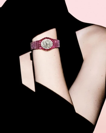 Close-up of a wrist wearing a luxury diamond and ruby wristwatch against a dark garment and pink backdrop