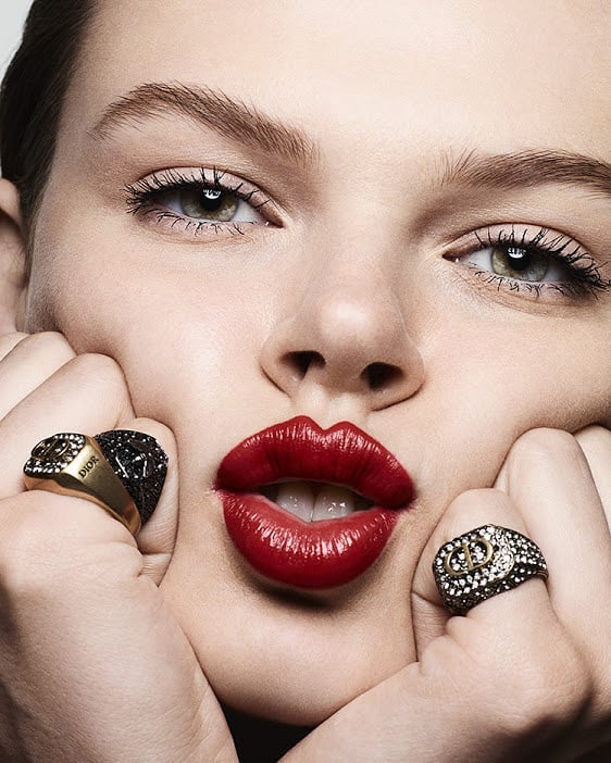 Close-up of a woman with bright red lipstick and several rings on her fingers posing indoors, focusing on beauty and fashion.