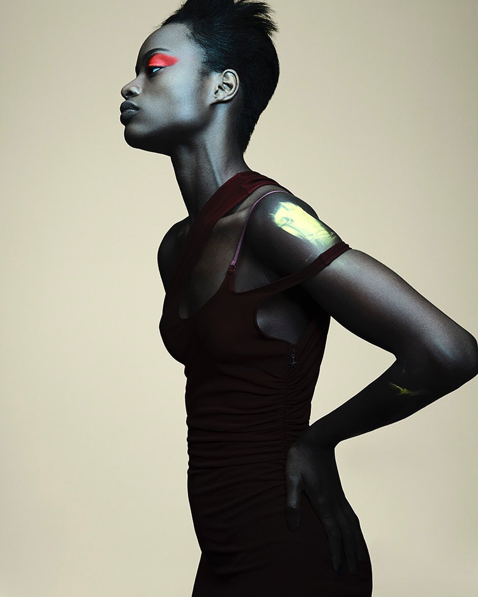 Elegant portrait of a Black woman in a burgundy dress with makeup, facing left against a neutral backdrop in a studio setting.