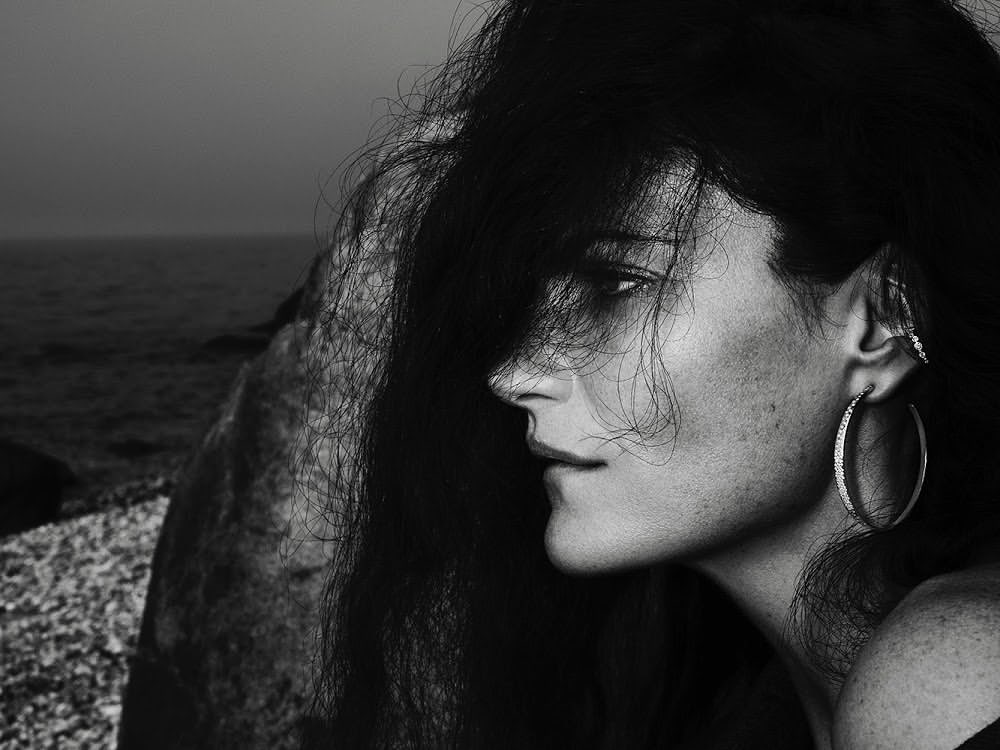 Close-up profile of a woman with dark hair covering her face, wearing a hoop earring against a rocky shore background