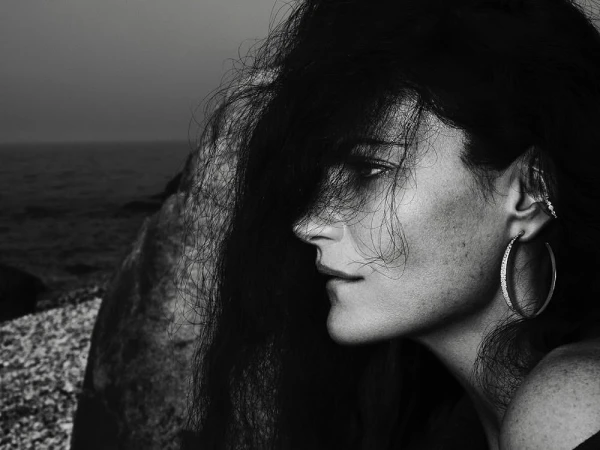 Close-up profile of a woman with dark hair covering her face, wearing a hoop earring against a rocky shore background