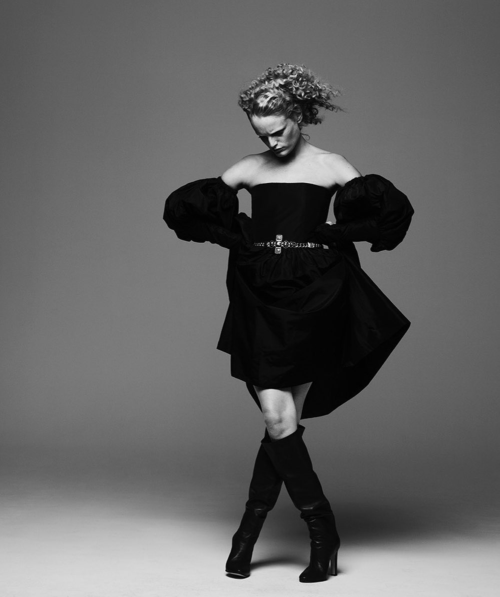 Monochromatic fashion portrait of a woman in a black dress with voluminous sleeves and knee-high boots in a studio setting.