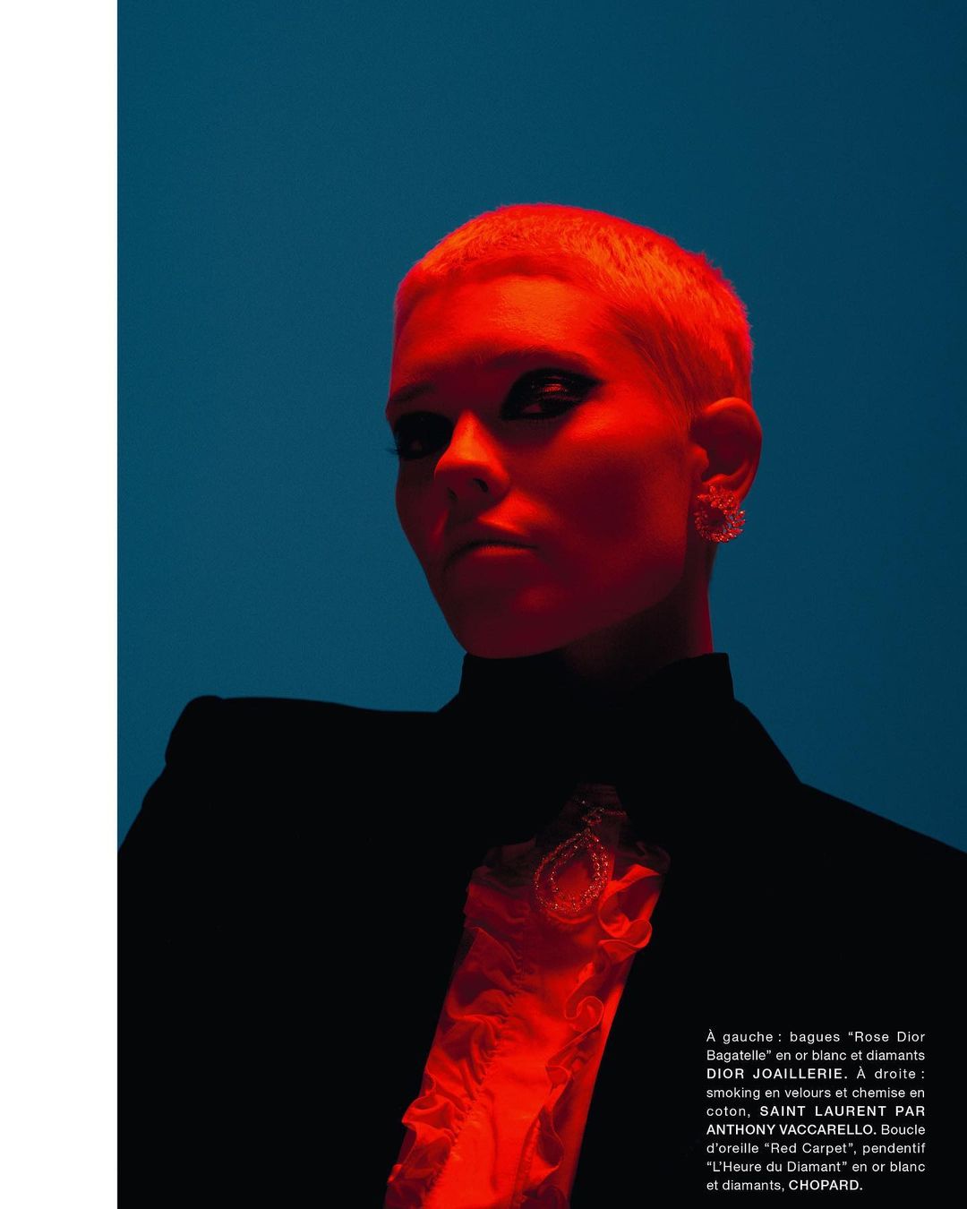 Glamour shot of a short-haired woman in a black blazer against a dark cyan background with red accent lighting and jewelry details.