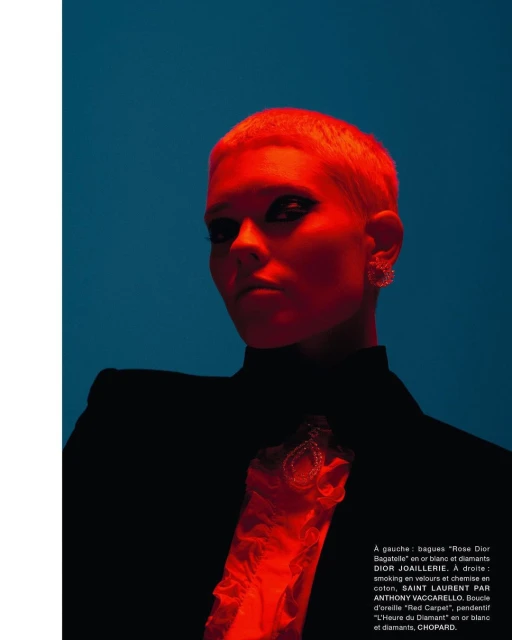 Glamour shot of a short-haired woman in a black blazer against a dark cyan background with red accent lighting and jewelry details.
