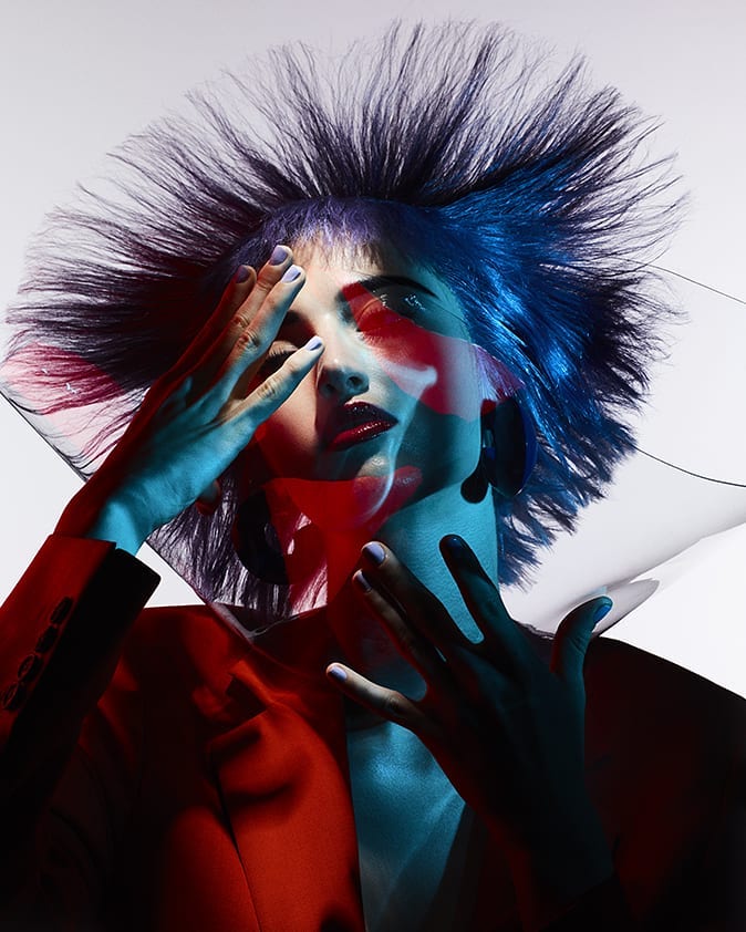 Glamour shot of a woman in a red jacket, with blue hair and hands near her face, illuminated with red and blue lighting.