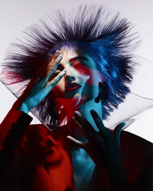 Glamour shot of a woman in a red jacket, with blue hair and hands near her face, illuminated with red and blue lighting.