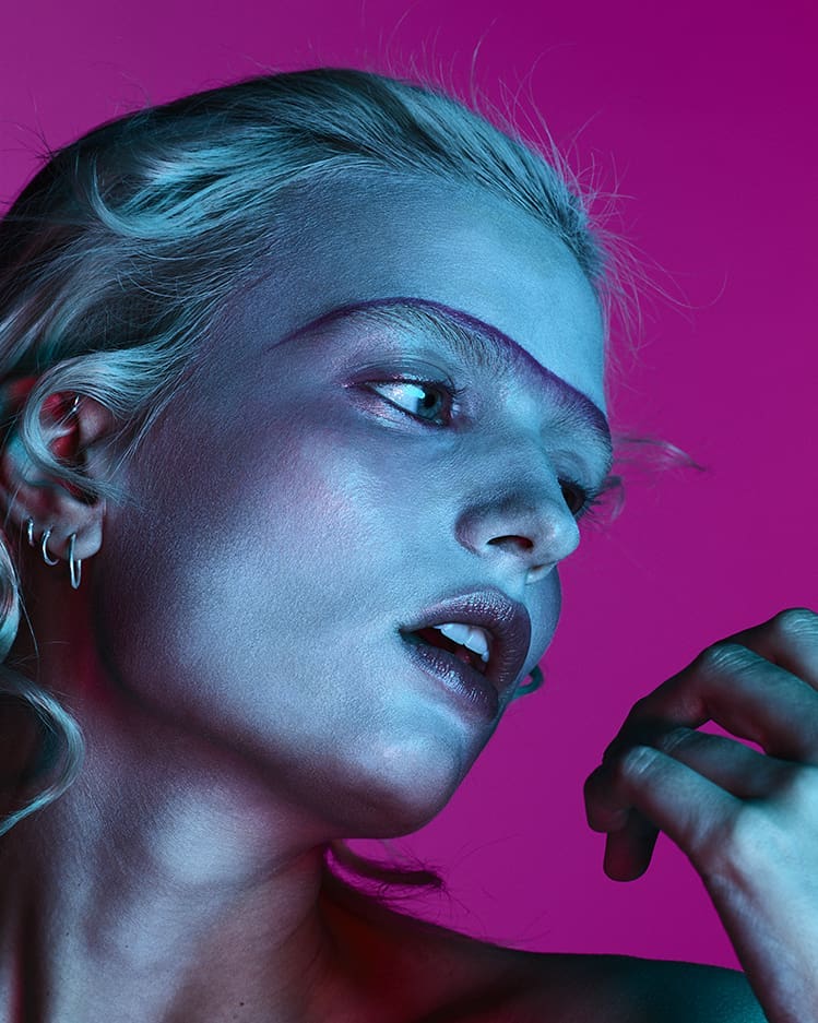 Close-up portrait of a young woman with light hair and purple accents against a pink background, illuminated with cool blue lighting.
