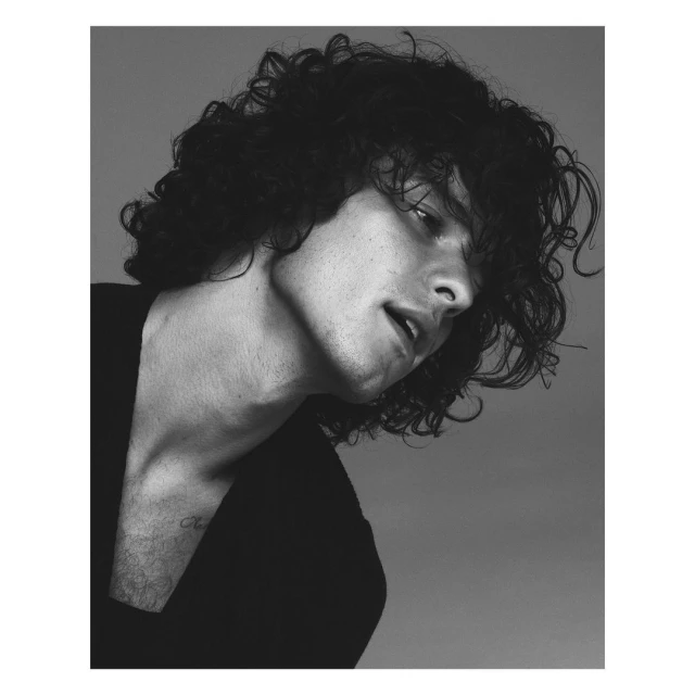 Monochrome portrait of a handsome adult male with curly dark hair, wearing a black shirt against a grey seamless background.