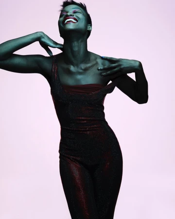 Model with short dark hair in a shimmering dark jumpsuit posing against a pale background