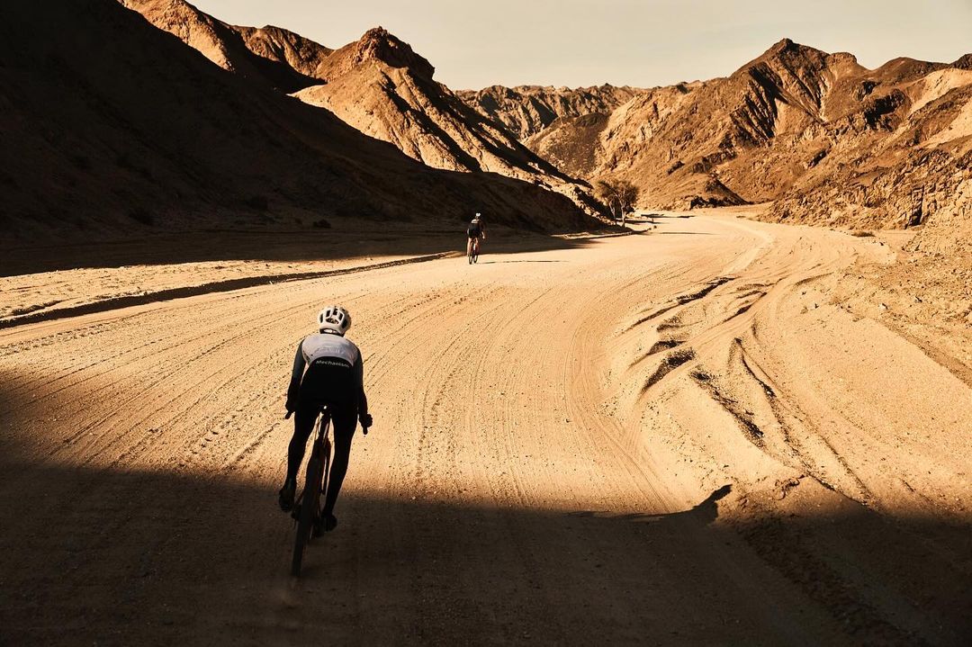 Two cyclists ride through a desert landscape with mountains in the distance, bathed in warm light with shadows creating contrast.