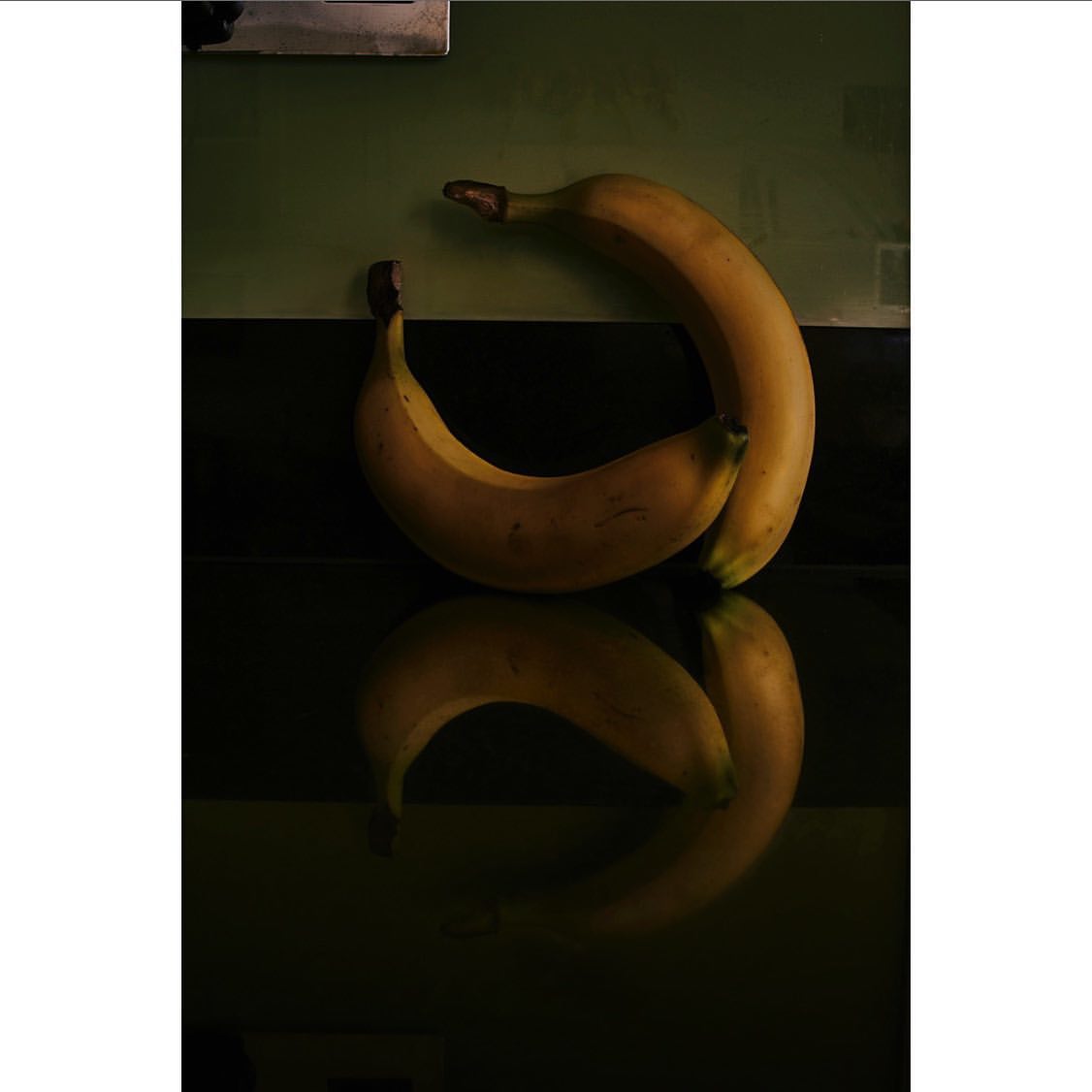 Still life shot of two ripe yellow bananas reflecting on a dark surface against a muted green background, suggesting a food photography theme.