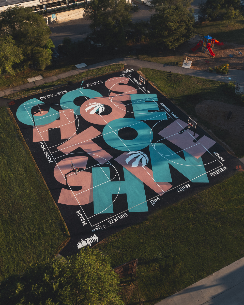 Aerial view of a colourful basketball court with large lettering and basketballs painted on it, surrounded by green grass and trees.