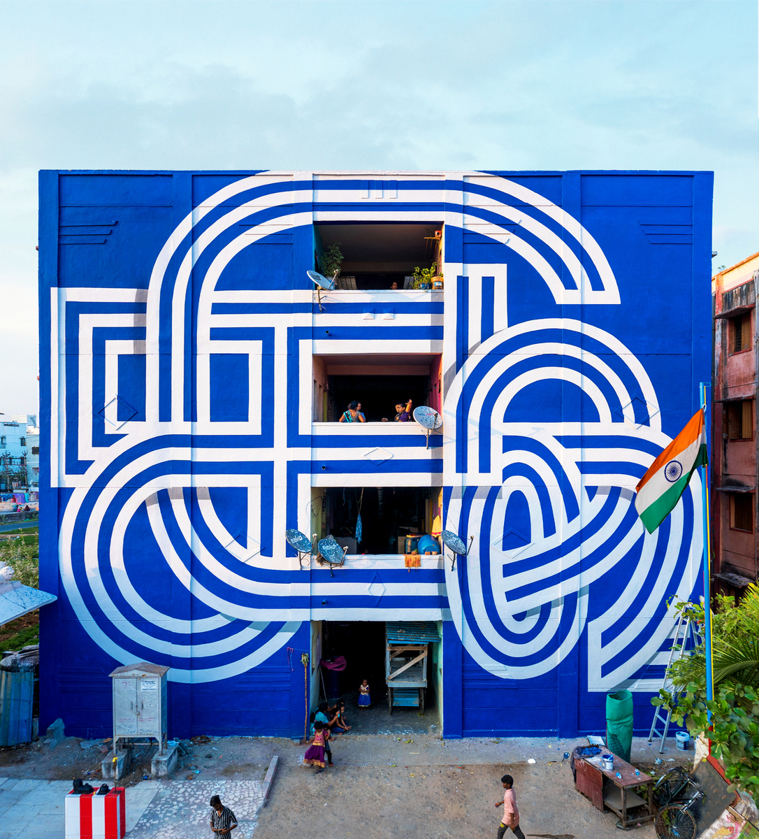 Blue building with white abstract mural art, balconies, people, and an Indian flag on a clear day in an urban outdoor setting.