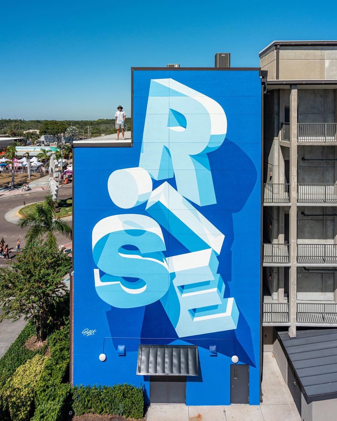 Large blue mural on building features the word 'RISE' in bold 3D letters, with a person standing on the edge of the rooftop.