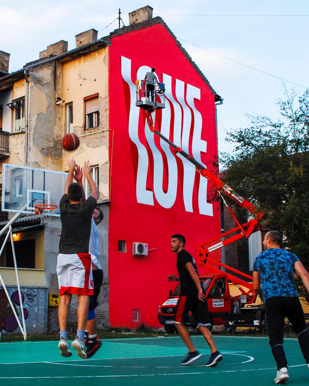 Outdoor basketball game on an urban court with a red building mural that says LOVE being painted by a worker in a crane basket.