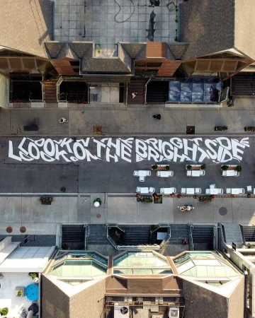 Aerial view of a street with the text "LOOK ON THE BRIGHT SIDE" painted on the ground, surrounded by buildings and planters.