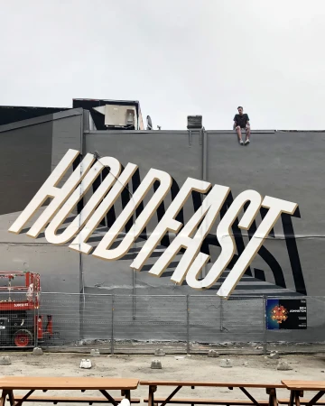 Outdoor mural featuring the word HOLDFAST with an adult male sitting on top of the building with tables in the foreground.