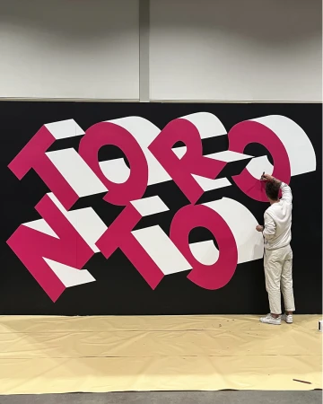 Adult male painting dimensional graffiti art of 'Toronto' in pink and white on a black wall indoors, wearing a white outfit.