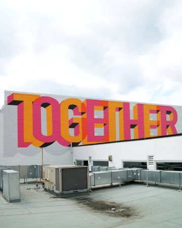 Eye-level shot of the word 'TOGETHER' in orange and pink, painted on the side of a white building with rooftop HVAC systems below.