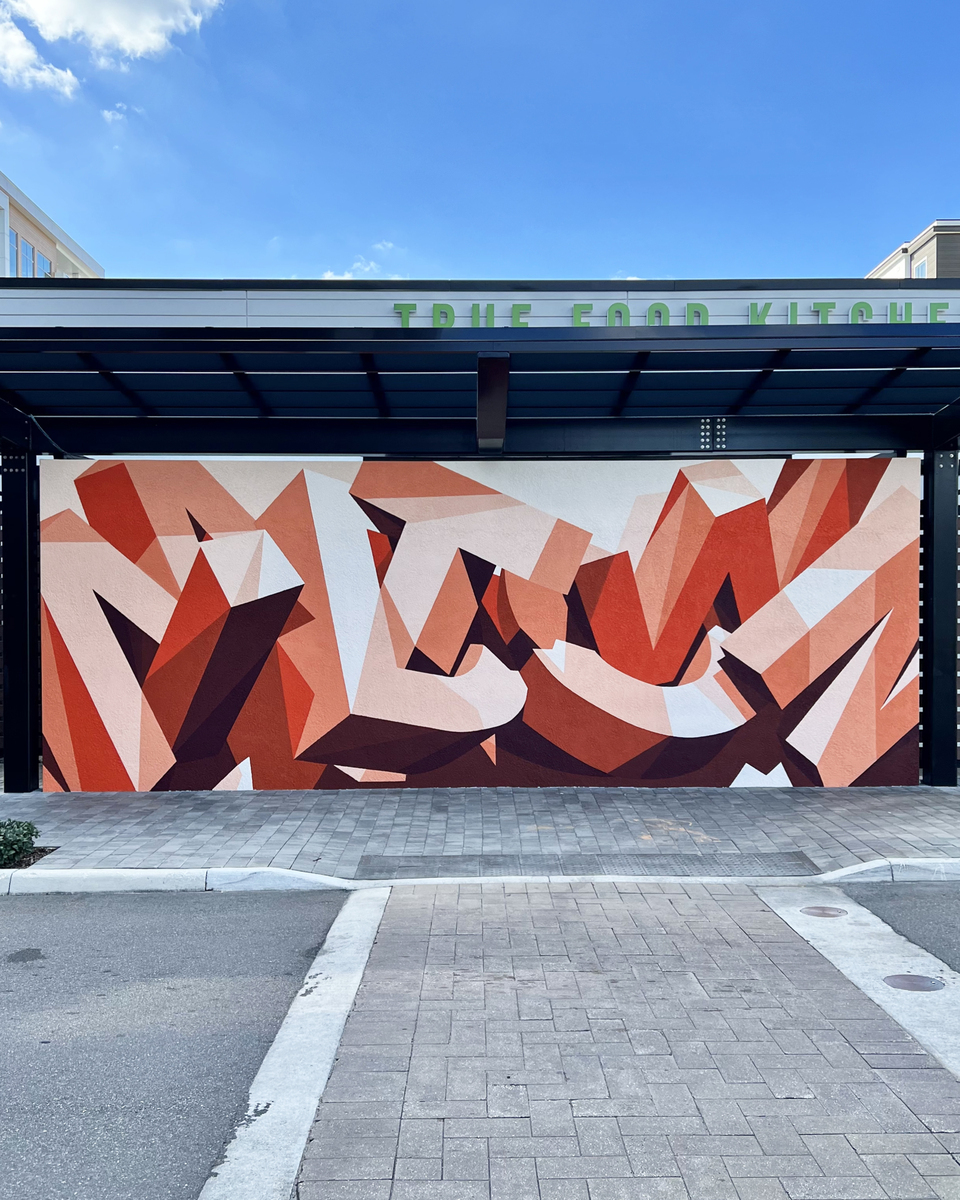 Exterior shot of an abstract geometric mural featuring shades of red, orange, and white painted on a wall beneath a dark metal awning.