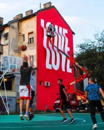 Men playing basketball on an outdoor court in front of a red building wall painted with large white typography by Ben Johnston