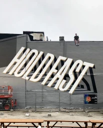Large 3D typographic mural reading HOLD FAST on a grey exterior wall with a person sitting on the roof edge above.