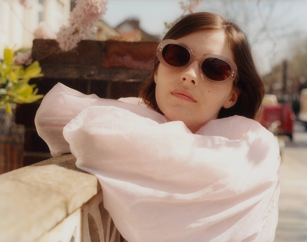 A woman with sunglasses and a pink blouse poses outdoors near greenery on a sunny day, captured in a medium shot.