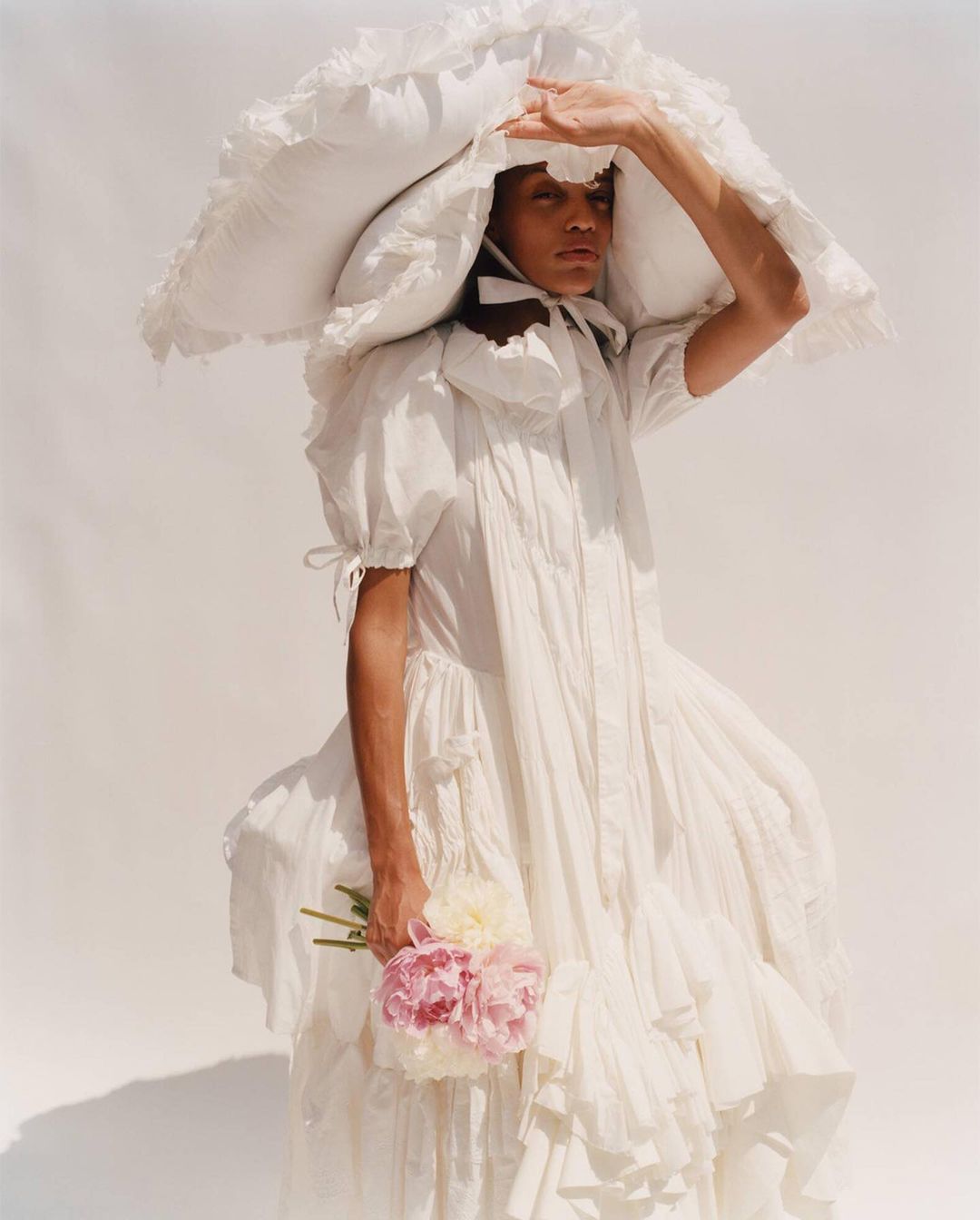 Person in ruffled white dress and oversized hat shields their eyes while holding flowers against a plain white background.
